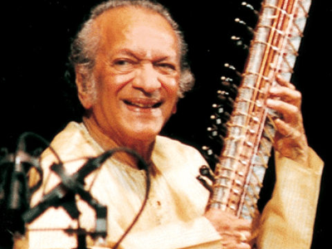 Portrait photograph of musician Ravi Shankar wearing a cream top and playing an instrument.